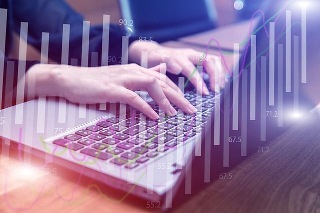 Tuvplāns ar rokām, kas raksta uz portatīvā datora tastatūras, virs kurām uzmontēti caurspīdīgi finanšu stabiņu un līniju grafiki.
